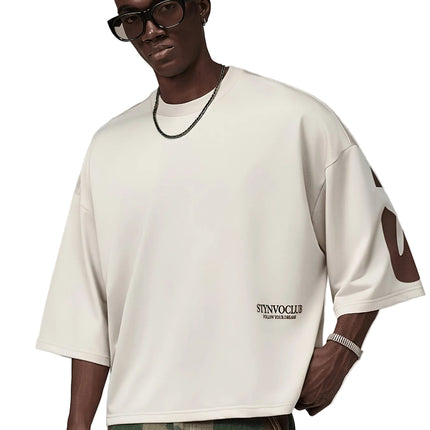 Oversized heren T-shirt in khaki, voorkant met zwarte STYNVOCLUB-letterprint, ronde hals, verlaagde schouders en grafische mouwprint op witte achtergrond.