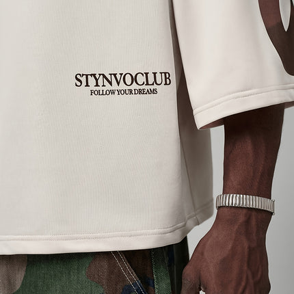 Oversized heren T-shirt in khaki, voorkant met zwarte STYNVOCLUB-letterprint, ronde hals, verlaagde schouders en grafische mouwprint op witte achtergrond.