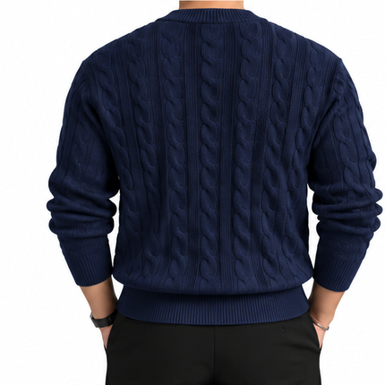 Heren kabeltrui met ronde hals, lange mouwen en klassiek kabelgebreid patroon, gedragen door een mannelijk model met een witte pantalon tegen een witte achtergrond.