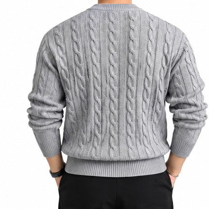 Heren kabeltrui met ronde hals, lange mouwen en klassiek kabelgebreid patroon, gedragen door een mannelijk model met een witte pantalon tegen een witte achtergrond.